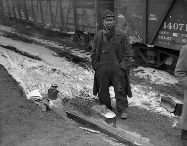 A man (maybe a railroad worker or a tramp) standing along a railroad track next to a small cooking fire. Railroad cars are in the background.