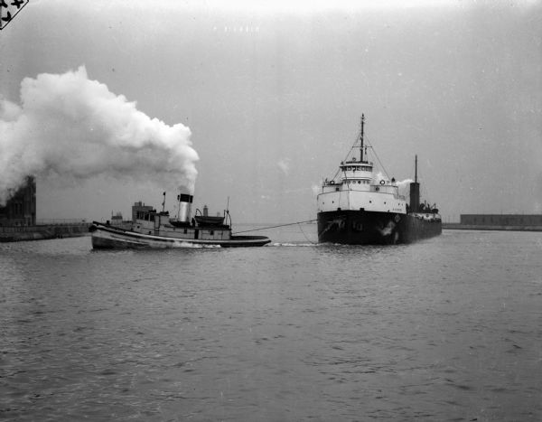 A tugboat pulling a steamer in the harbor of Lake Michigan. Steam is billowing from the stacks of the steamer.