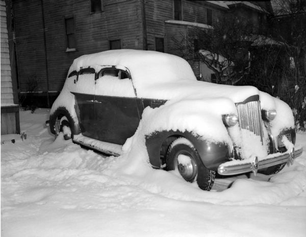 Winter scene with a car buried in fresh snow while parked outside a building.