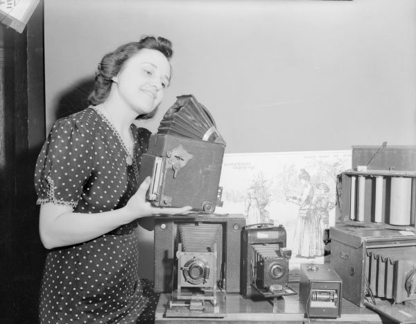 Woman posing holding a camera near a table full of old cameras.