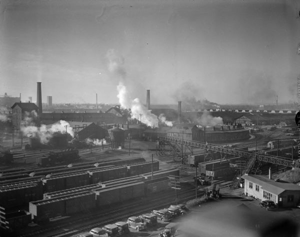 Elevated view of West Milwaukee shops of the Chicago, Milwaukee and St. Paul railroad. Railroad train cars are stopped on the tracks and smoke is rising from the buildings in the background near smokestacks.