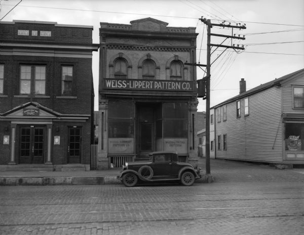 Exterior view of the Weiss-Lippert Pattern Company. A car is parked outside near the curb on the cobblestone street, and a chapel stands to the left.