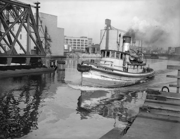 "Conrad Starke" (possibly a tugboat) cruising on the water near a bridge in an industrial area. The waterway is probably connected to Lake Michigan. Three men are standing on the boat.