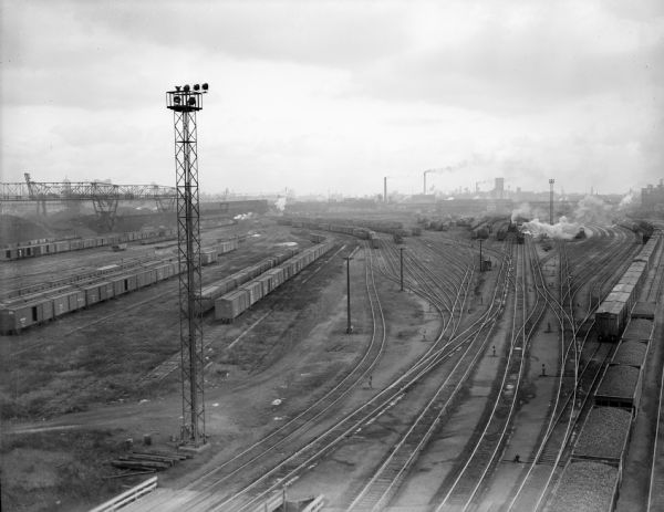 Elevated view of railroad yards with trains sitting on many of the tracks, though other tracks are sitting empty. Steam is rising from trains in the background, as well as from smokestacks in the distance.