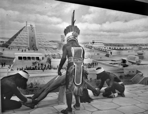 Diorama of Aztec sacrificial rites at the Milwaukee Public Museum. Two men are holding the man to be sacrificed in a splayed position while a third man stands above him. A backdrop of an Aztec city appears behind the men, filled with a crowd of people watching.