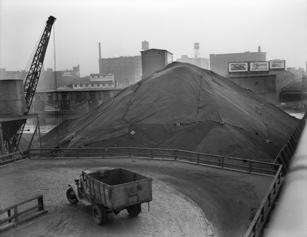 Elevated view of a large pile of coal sitting under a tarp, with an empty truck waiting nearby. Industrial buildings and water towers are across the river. One building has a sign that reads: "United State Gypsum Co."
