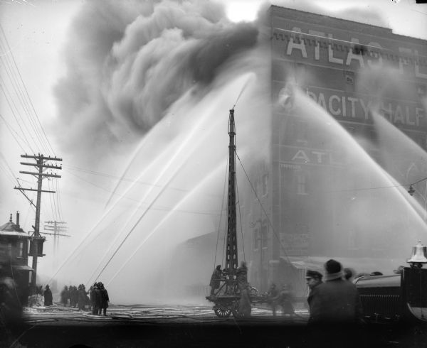 Fire at the elevator of the Atlas Flour Mill.  Two men are working a water hose tower in the center, and fire fighters are aiming jets of water at the building from many sides.  Smoke is billowing out in the upper left area of the image.