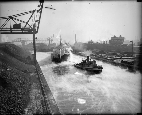 Elevated view of tugboat pulling a steamer caked in ice in a canal. Steam is rising from the water and an open drawbridge can be seen in the background. On the left side are large piles of coal or coke.