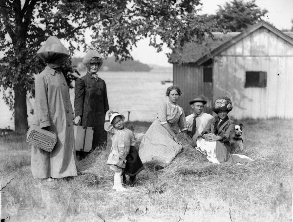 A group of people under a tree in a farmyard, with possibly a lake in the background. Two women, with heavy netting on their hats, hold luggage. A toddler is standing in the center, while two women, a man, and a dog are sitting on and around a pile of hay on the right.