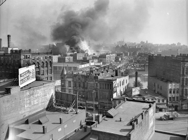 Elevated view across rooftops towards smoke rising from a burning building in the city. The building on the left has a billboard supporting the Packers. A bridge and river are in the center, and a wide boulevard is in the distance.
