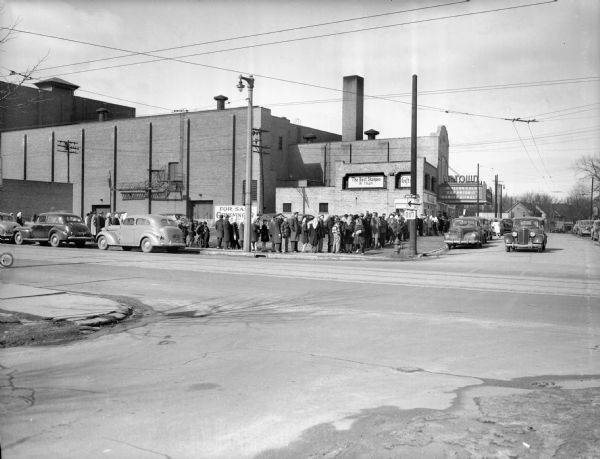 View across intersection towards a line of people formed down and around a street corner at the Uptown Theatre. The marquee advertises showings of "San Antonio" and "Danger Signal," both in technicolor. Street signs and other advertisements are on buildings and the street corner. Cars are parked along the street.