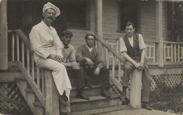 Four men are sitting on porch stairs and railing posts. The man on the left is wearing a chef jacket and hat, and also an apron.