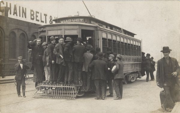 Men piling into an electric trolley with a sign that reads: "Viaduct". The streetcar has grating on the end, which several men are standing on. A sign on a building in the background reads, in part: "Chain Belt &".
