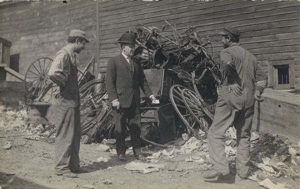 Three men standing in a junkyard with piles of wheels, chairs, and other wooden items in the background near the side of a building. The ground is littered with papers. The man in the middle is wearing a suit and hat, while the other two men are wearing overalls and caps.