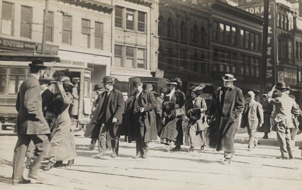 A group of well-dressed men and women are walking across a street. They are all wearing hats and a couple of the woman are carrying fur muffs. A streetcar and an automobile are in the background.