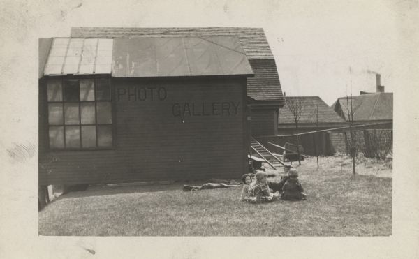 A group of four young children sitting in a circle on the grass. Several buildings are in the background, and a few bare trees are on the right. The building in the background has a large window and what may be a skylight. A sign near the window reads: "Photo Gallery".