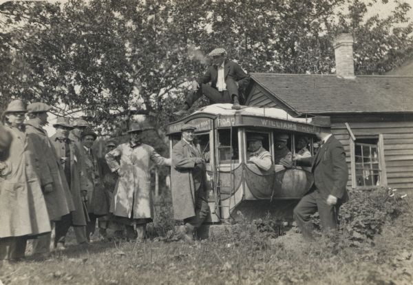 A group of men are gathered around a stagecoach which is mounted on a platform in a yard, with buildings in the background. Some men are sitting inside, while others are standing around in a semicircle. A man is sitting on the roof of the vehicle smoking. "Rail Road," "A Steam Boat Line," and "Williams House" is written on the exterior of the carriage.