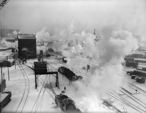Elevated view of railroad yard, with engines and cars on some of the tracks. Smoke is billowing out from many of the engines, covering much of the yard. Buildings and rail equipment are on the left.