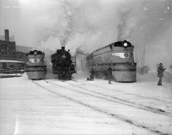 Front view of three trains in a snow-filled train yard. The middle train is an old-fashioned steam engine flanked by more modern trains. Three men are standing nearby.
