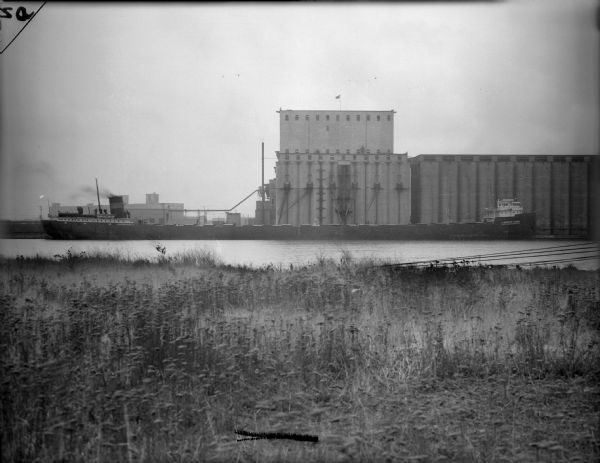 View from shoreline of a grain ship on the Kinnickinnic River. There are industrial buildings in the background.