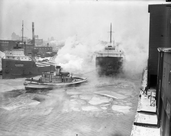 Tugboat pulling freighter "Mariposa" on Kinnickinnic River. The river has large chunks of ice floating on top, and industrial buildings are in the background.