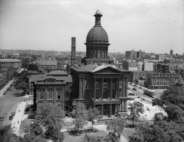 Elevated view of Milwaukee County Court House, with a tree-filled park area in the foreground and the city in the background. There is debris on the roof, and three men are working on the top of the courthouse dome.