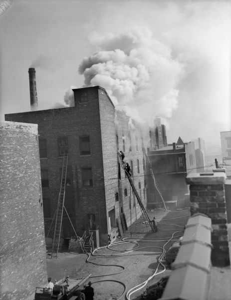 Elevated view from rooftop of smoke billowing out of the roof of a burning brick building. Fire fighters are climbing on ladders on the side of the building, and water hoses snake along the ground.