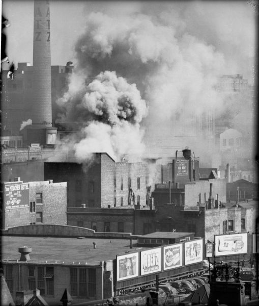 Elevated view from a distance of a fire. A large cloud of smoke is billowing out of the burning building and fire fighters are climbing on a ladder. A building in the foreground has several billboards. A smokestack in the background has a sign that reads: "Blatz".