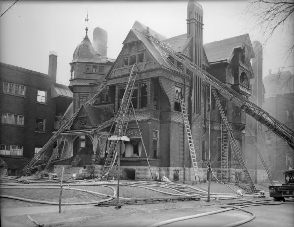 A large Victorian-style house gutted by a fire. Ladders are leaning up on the sides, reaching up many different levels. Two people are on the roof spraying water, and hoses are spread over the lawn. Icicles are hanging from the ladder rungs and parts of the exterior. A fire engine is parked on the street on the right side. One of the street signs reads: "W. Wisconsin Avenue".