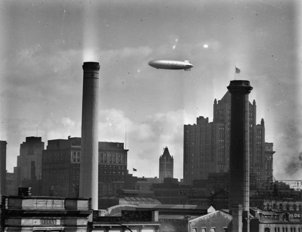Blimp (dirigible) flying over downtown buildings. There are two smokestacks in the foreground, and on the right, a sign, from the back, for "The Electric Co."