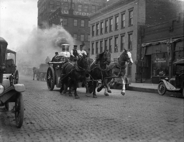 Three men in a horse-drawn fire engine racing down a downtown street.