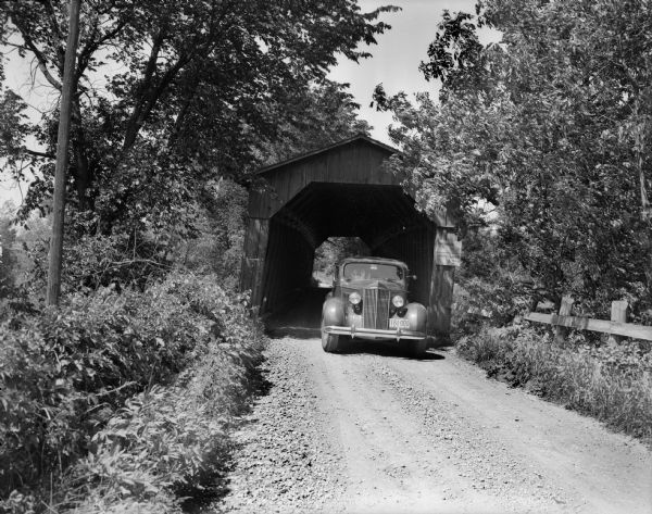 A car is parked at the entrance of a covered bridge. There is a fence on the right side of the road. Sign on the bridge reads: "Bridge No.7, Safe Load 3 ton."
