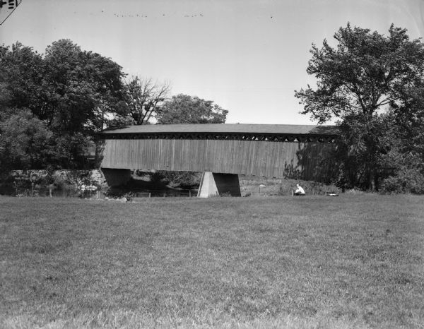 A man in the distance is sitting in a field, with a covered bridge behind him spanning a river. Trees are at each end of the bridge, and a fence runs along the shoreline on the left.