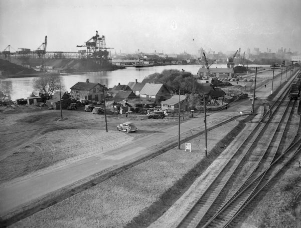 Elevated view of Jones Island, with some houses near the water and an industrial area across the river. On the right are a number of railroad tracks next to a road. A sign in the foreground reads: "USA WORK PROGRAM WPA."