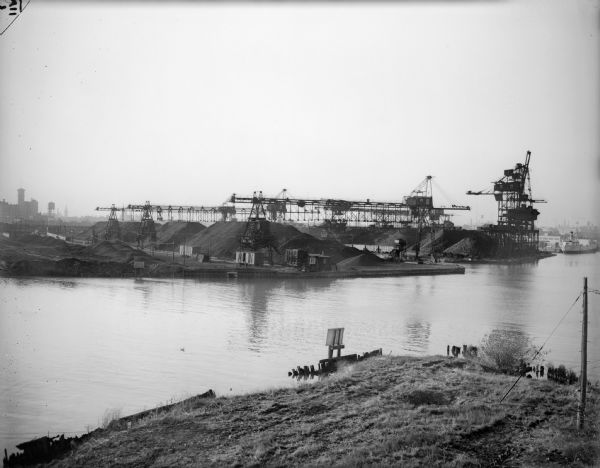 From Jones Island, looking across to a manufacturing area with cranes and large piles of coal or coke? In the lower foreground is a bare hill, with old pilings in the water and signs at the water's edge on both side of the bay.