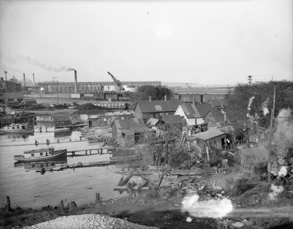 Elevated view of bay shoreline, with dwellings along the bank. A few boats are in the river. In the background is a factory with a smokestack and railroad cars on a track.