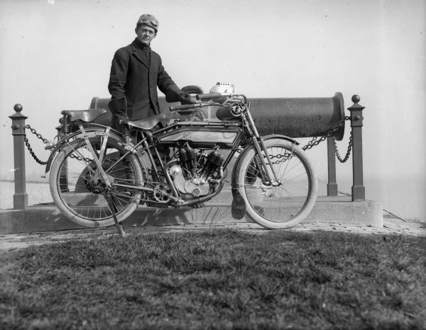 The photographer J. Robert Taylor posing outdoors with his motorcycle in front of a large cannon. The cannon is mounted on a low pedestal and is enclosed by a fence and chains.