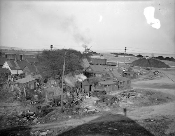 Elevated view of a cluster of old dwellings in the center, with broken shacks and rusted cars. In the background are railroad cars, and an industrial area with men working. In the distance is the shoreline of Lake Michigan. In the foreground is the shadow of a hill with a figure on top, presumably the photographer.