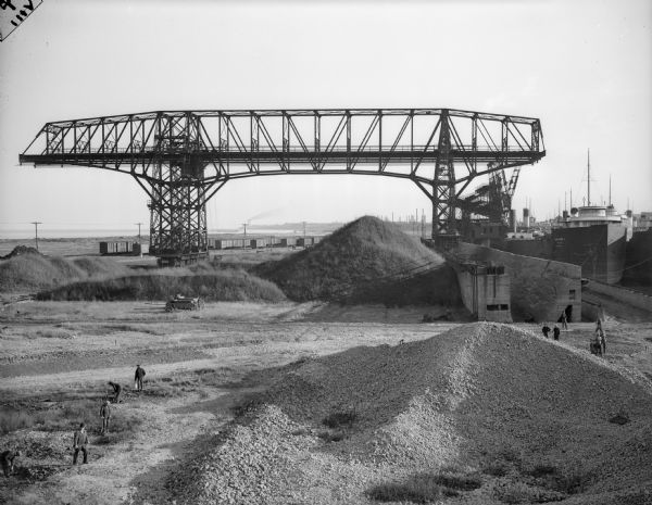 Men working near large dredge on Jones Island. Large piles of material are on the ground below the dredge.  There is a ship on the right. In the distance are railroad cars on tracks, and the shoreline of Lake Michigan.