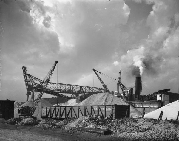 View across industrial yard towards piles of sand around a dredge. There is a railroad car and crane in front of the dredge. In the background is a large smokestack on the ship which is carrying the dredge.