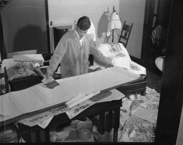 Slightly elevated view of a young man preparing a piece of wallpaper to be hung. On the ground and under furniture newspapers have been laid to protect the room.