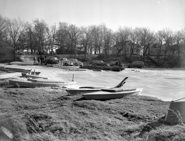 Frozen river shoreline, with boats sitting on the grassy banks in the foreground. Across the icy shoreline are cars parked near the boat loading area, with men standing nearby. Houses line the hill on the right. One of the vehicles ha a sign that reads: "Waukesha Co. Ambulance" on its side.