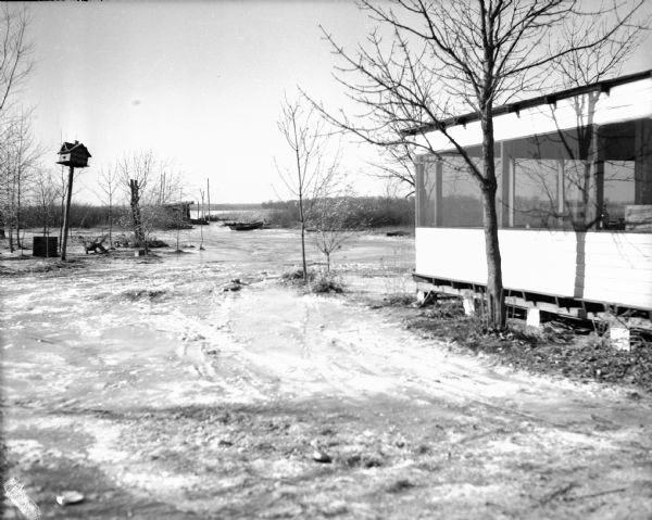 Ground around lodge covered in mud and ice, some from the frozen river in the background. There is a large birdhouse on a pole on the left.