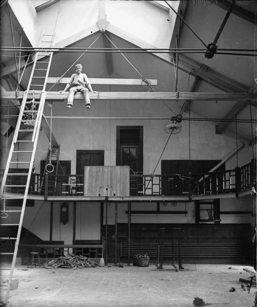 A man is sitting high up on a rafter in a building with a skylight. There is a ladder to his left, and a balcony is behind him.