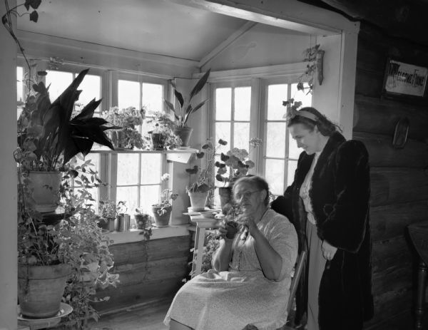 Two women in an alcove filled with potted flowers and plants. The older woman is sitting and the other woman, wearing a fur coat, is standing behind her.