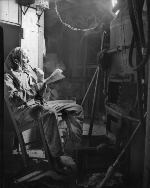 Taylor is relaxing, sitting in a chair tilted back, and smoking his pipe and reading his paper in front of an open heater. A kerchief is covering his head. Smoke is floating around the light in the upper right.