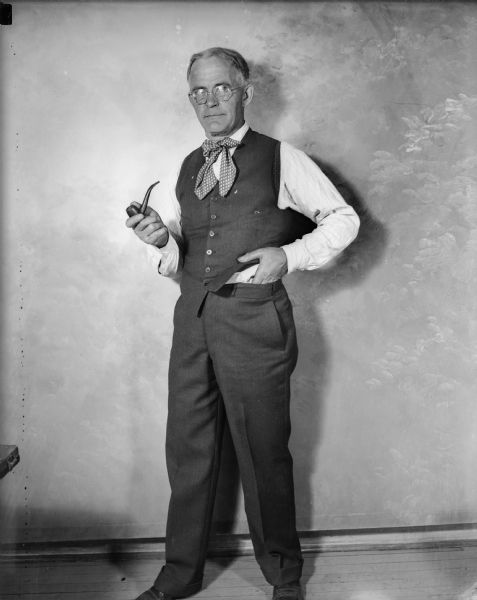 J. Robert Taylor, the first photographer for the <i>Milwaukee Journal</i>, poses in a formal portrait, standing with his pipe. He wears a cravat style tie and a vest.