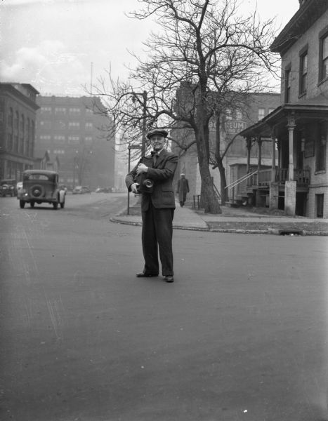 J. Robert Taylor standing in the middle of a street, holding a large camera. On the left, a car is driving by, and a person is walking on the sidewalk in the background.