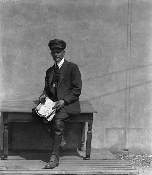 Self-portrait of the photographer sitting on a table near a wall wearing a hat and holding a newspaper and pipe.