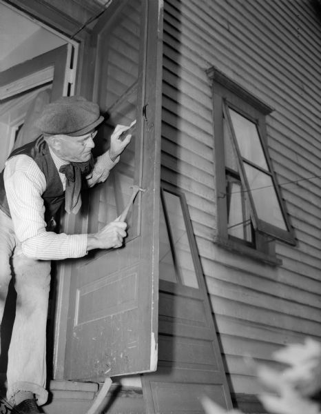 J. Robert Taylor leaning out of a doorway, fixing a screen door with a hammer in his right hand.  Another door is leaning against the house to the left of an open window.